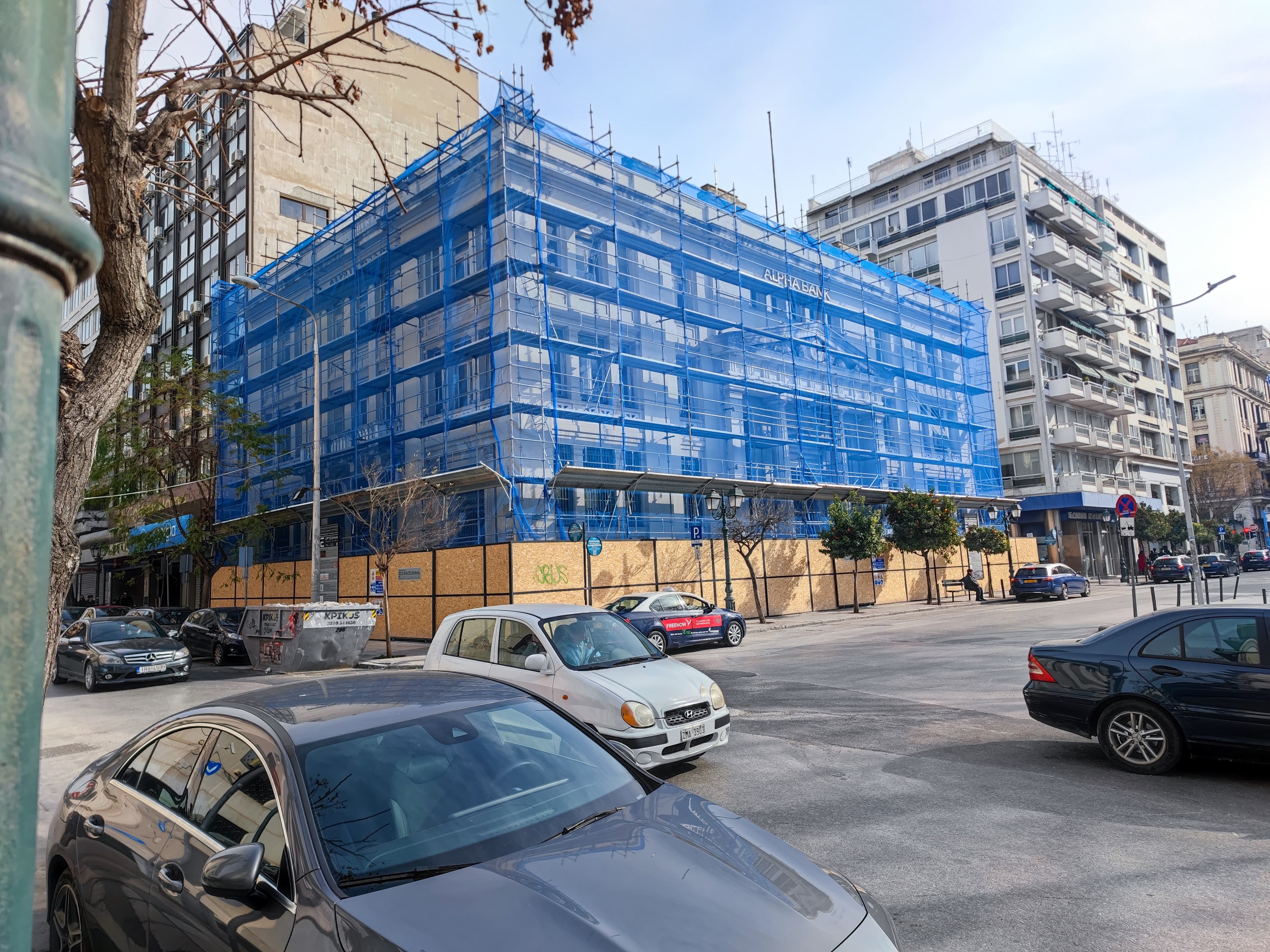 Alpha Bank building wrapped in blue scaffolding netting on a city street corner with cars.